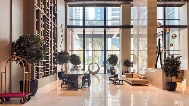 A spacious lobby with a high ceiling, large windows, and modern furnishings including a sofa, chairs, and a bar area.