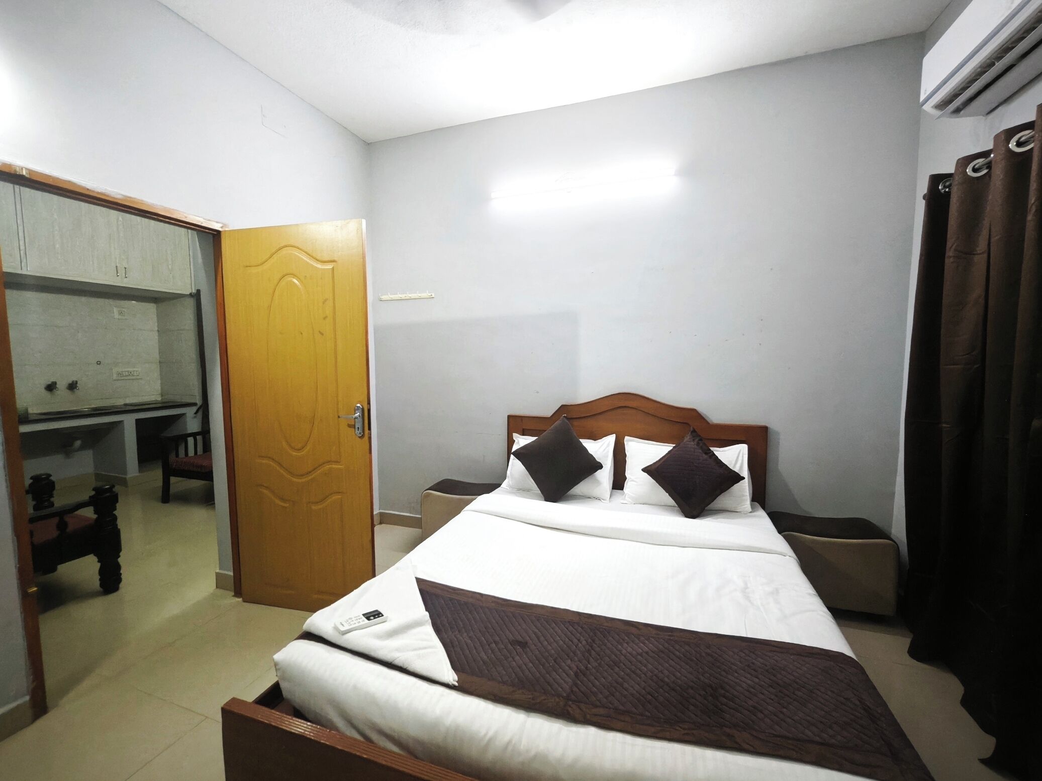 A hotel room with a bed, bedside tables, a desk with a chair, a wardrobe, and an air conditioning unit.