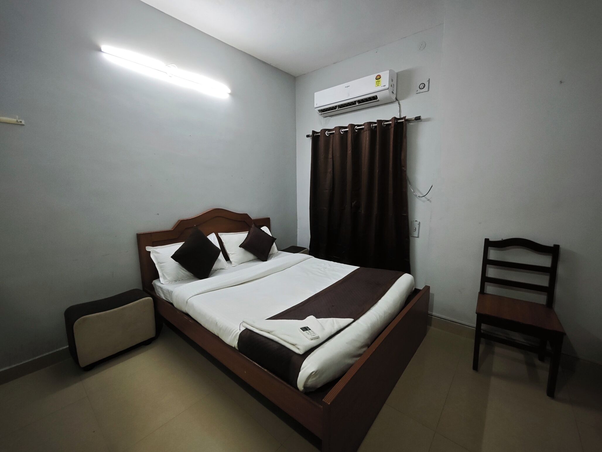 A neatly made bed with a wooden headboard, a bedside table, a chair, a wall-mounted air conditioner, and a ceiling light.