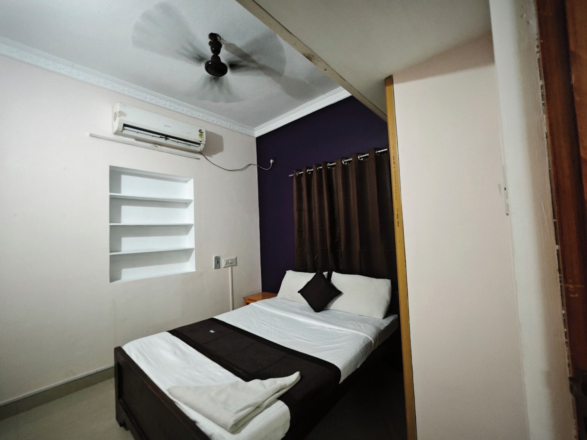 A small, single-bed room with a ceiling fan, an air conditioning unit, a window with blinds, and a dark-colored wall.