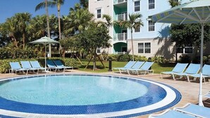 Una piscina circular con sillones de playa y sombrillas, rodeada de palmeras y un edificio con balcones.