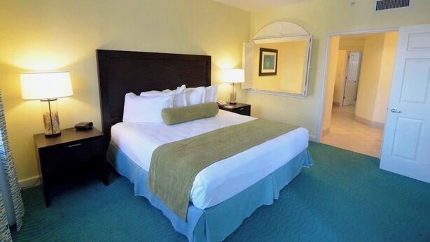 Una habitación de hotel con una cama grande, dos mesitas de noche con lámparas, una alfombra verde y una puerta que da a otra habitación.