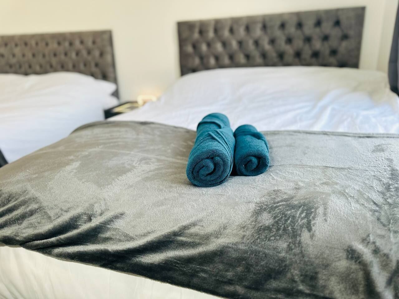 A bed with a grey upholstered headboard and pillows, and two rolled blue towels placed on the bed.