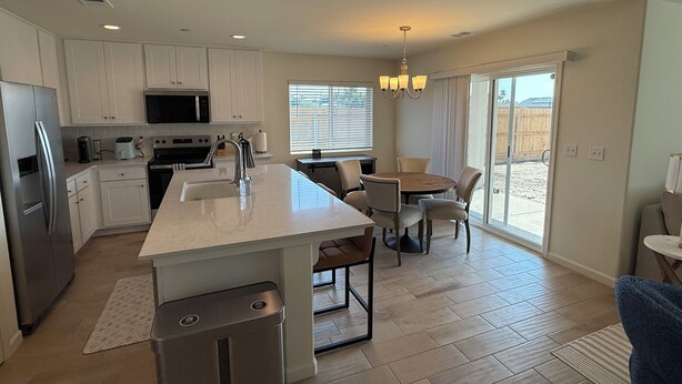 A modern kitchen with white cabinets, a stainless steel refrigerator, and a central island with a sink. There is a dining area with a table and chairs, and a sliding glass door leading to a patio. - New-construction, Pet-Friendly 2-bedroom house in Hanford (Hanford)