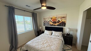 A bedroom with a bed, a ceiling fan, a window with curtains, a painting on the wall, and a nightstand with a lamp. - New-construction, Pet-Friendly 2-bedroom house in Hanford (Hanford)