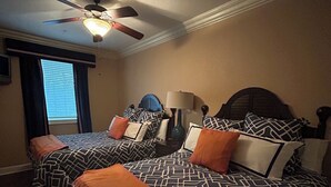 A bedroom with two beds, a ceiling fan, a window with blinds, and a lamp on a bedside table. -  3-BR Condo Auburn, AL  Walking distance and minutes from downtown and campus (Auburn)