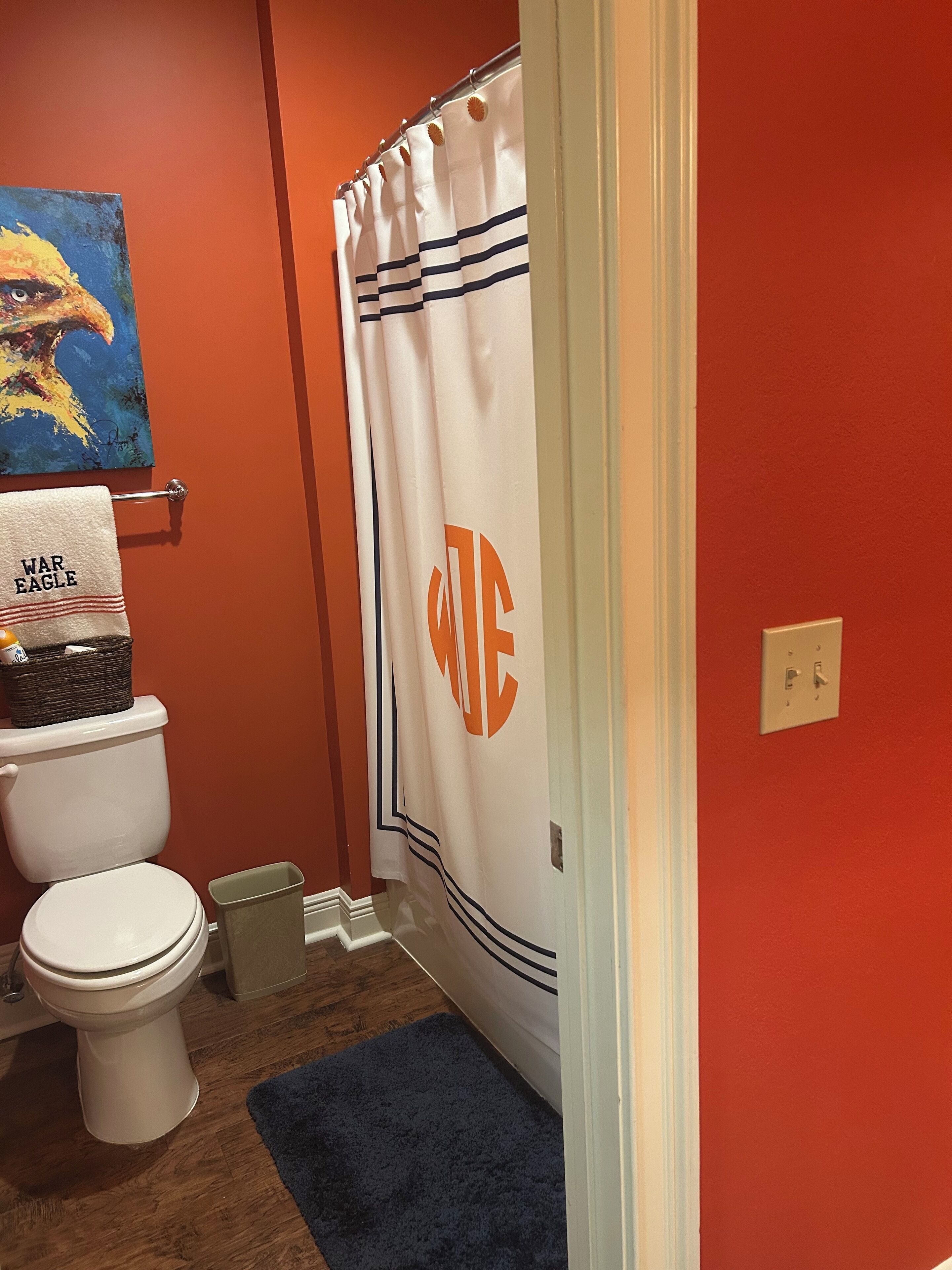 Ein Badezimmer mit einer weißen Toilette, einem blauen Teppich, einem weißen Duschvorhang mit orangefarbenen Lettern und einem Bild an der Wand.