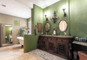 Ein Badezimmer mit freistehender Badewanne, einem Holzkosmetikschrank mit mehreren Schubladen, zwei Wandspiegeln und einer Zimmerpflanze.