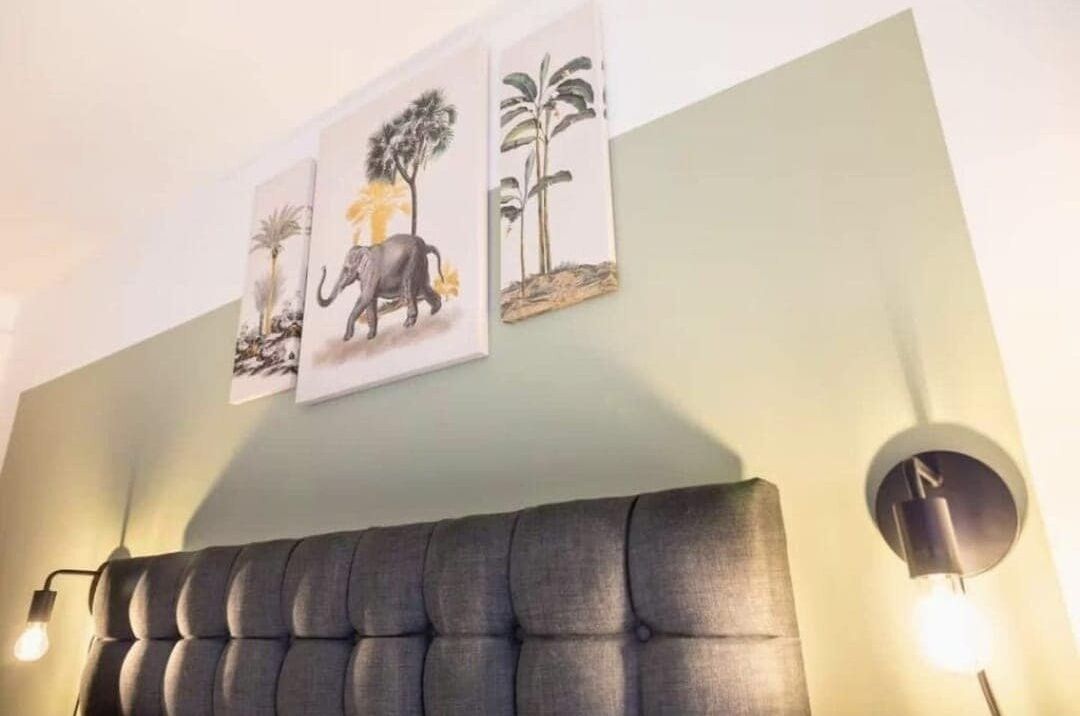 Una habitación con un cabecero tapizado en gris, una obra de arte en la pared que muestra elefantes y árboles, y una lámpara de pared.
