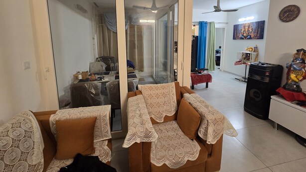 Una sala de estar con un sofá cubierto de encaje, una mesa de centro y un televisor. Hay un ventilador de techo y una estatua sobre una mesa.