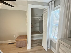 A room with a wooden floor, a white bench, a wicker basket, a closet with shelves, and a window with blinds.