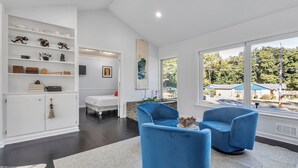 Een moderne woonkamer met een groot raam, een wit kastje met decoratieve objecten, en twee blauwe stoelen.