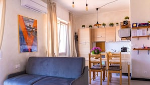 Una sala moderna con un sofá, un comedor con una mesa y sillas, una cocina con armarios de madera, y un estante en la pared con plantas y un reloj.