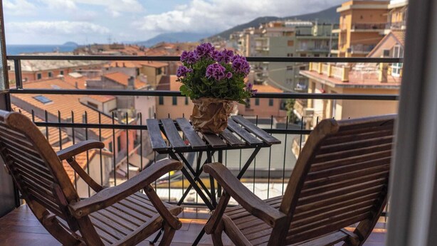 Un balcón con una mesa de madera y dos sillas, una planta en maceta con flores moradas, y vista a la ciudad.