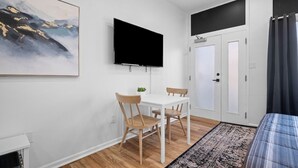Een moderne kamer met een witte muur, een wandgemonteerde flatscreen televisie, een witte tafel met twee stoelen, een zwart-wit tapijt met een patroon en een bed met een gestreept dekbed.