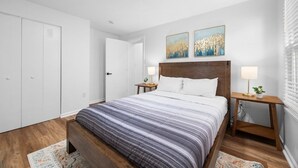 Un dormitorio con cama, dos mesitas de noche, una mesita auxiliar, cabecero de madera y una puerta.