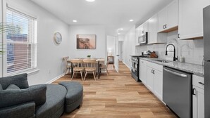 Una cocina moderna con armarios blancos, horno empotrado y lavavajillas. Hay una zona de comedor con mesa y sillas, y un sofá gris al frente.