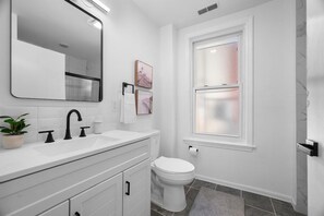 Un baño moderno con un lavamanos blanco, grifo negro y un espejo grande. Hay un inodoro y una ventana con vidrio esmerilado.