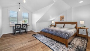A bedroom with a sloped ceiling, a wooden bed, two bedside tables with lamps, a desk with chairs, and a window with blinds.