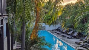 Un espace piscine aménagé avec des chaises longues, des palmiers et un panneau indiquant les heures d’ouverture de la piscine.