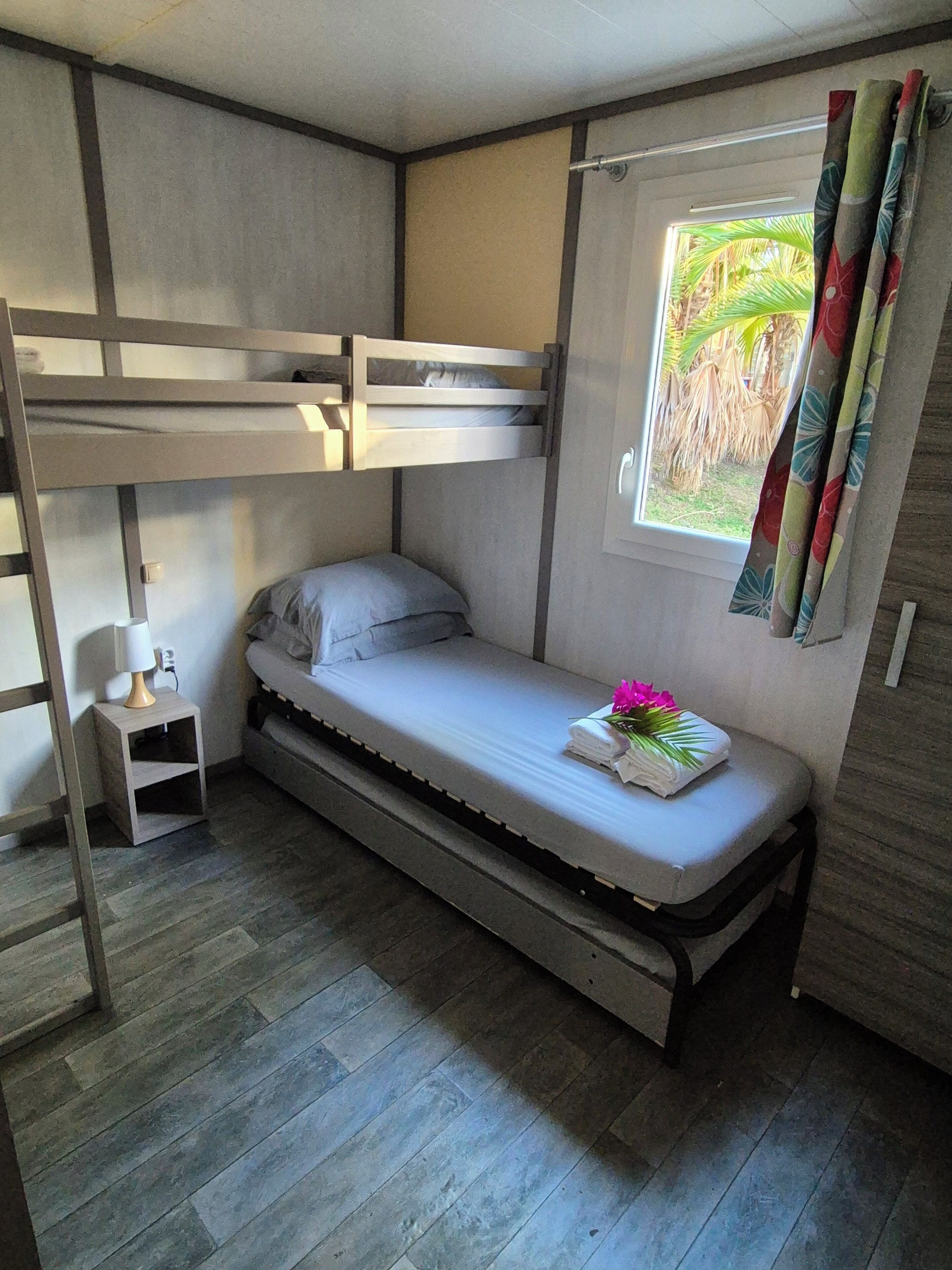 Habitació senzilla amb literes, una petita taula de nit amb làmpada i finestra amb cortines.