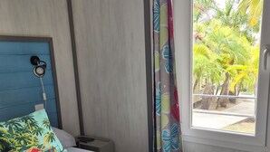 Une chambre d’hôtel avec un lit, un oreiller, une table de chevet, une fenêtre avec des rideaux et une vue sur les palmiers à l’extérieur.