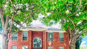 A red brick house with a central entrance, surrounded by trees and a pathway. - Pickleball + Pool + Bkyd Oasis + Centrally Located (Frisco)