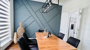A modern dining room with a wooden table and black chairs, a geometric-patterned wall, and a large window with blinds. - Pickleball + Pool + Bkyd Oasis + Centrally Located (Frisco)