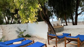 A beachside lounge area with wicker lounge chairs and blue cushions, a white wall, and a tree providing shade. - 5 BED Beach front Villa Paje (Paje)