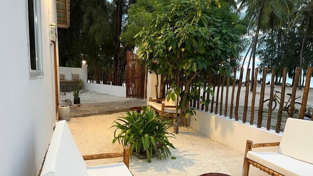 A covered outdoor seating area with white cushioned chairs and a small round table, surrounded by plants and a bamboo fence. - 5 BED Beach front Villa Paje (Paje)