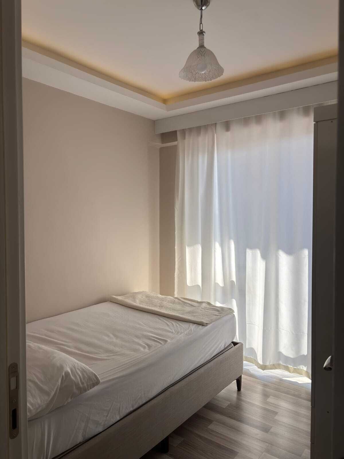 A bedroom with a single bed, a hanging light fixture, and a window with sheer curtains.