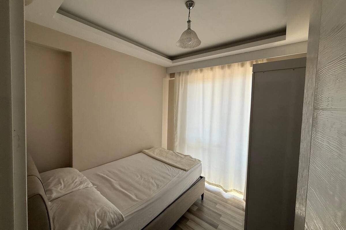 Habitació petita amb un llit individual, ventilador al sostre, finestra amb cortines transparents i una porta.
