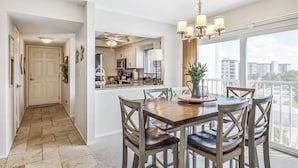 A dining area with a wooden table and chairs, a chandelier, and a view of the city through large windows. - Beautiful Sand Caper 408. 2B/2B condo steps from the Gulf. AMAZING Sunsets (Fort Myers Beach)