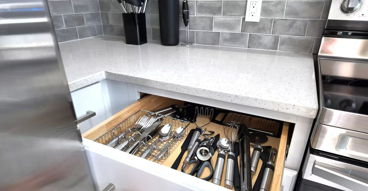 Uma cozinha com forno de aço inoxidável, bancada branca e organizador de gaveta com utensílios.