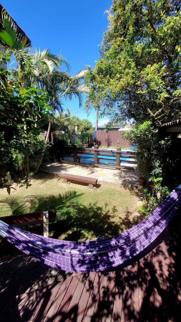 A backyard with a wooden deck, a hammock, a swimming pool, and various plants. - Pousada Uluwatu (Imbituba)