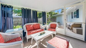 Uma varanda coberta com móveis de vime, uma mesa de vidro e vista para uma casa por uma porta aberta.