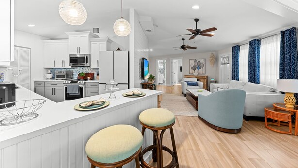 Uma cozinha moderna com armários brancos, uma ilha central com banquetas e uma área de jantar com sofá azul e uma luminária.