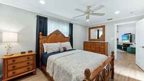 Quarto com cama de madeira, mesa de cabeceira, cômoda, ventilador de teto e televisão no quarto vizinho.