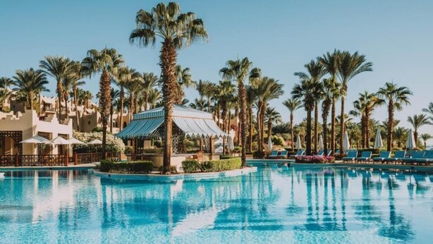 En pool vid ett semesterhotell, omgiven av palmer, med solstolar och en överbyggd paviljong.
