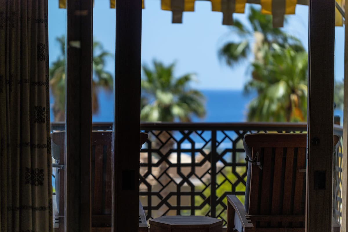 A wooden balcony with a lattice railing, overlooking a palm tree and the ocean.