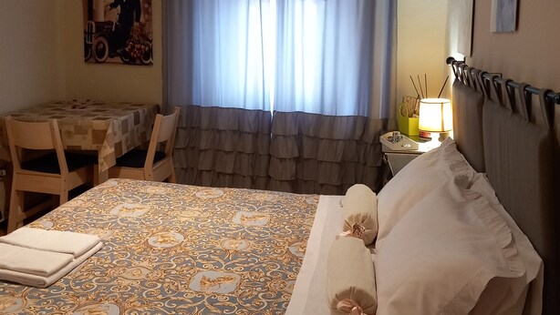 A room with a bed, a table, and a chair. There is a window with curtains, a wall-mounted air conditioner, and a lamp on a bedside table.