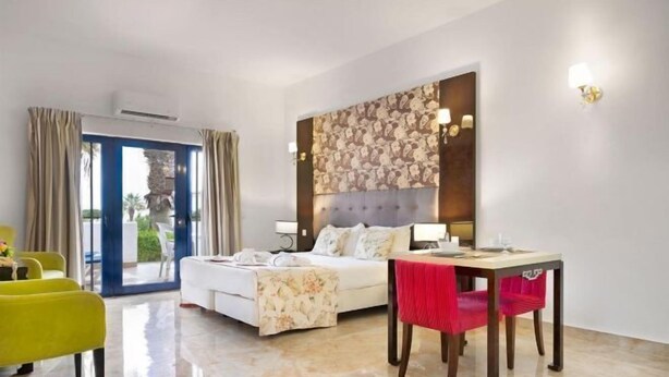 A hotel room with a bed, a dining table, a yellow armchair, and a view of the outdoors. - Grand Muthu Oura View Beach Club (Albufeira)