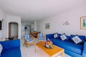 A living room with a blue sofa, striped chairs, a wooden coffee table, and a dining area with a table and chairs. - Grand Muthu Oura View Beach Club (Albufeira)