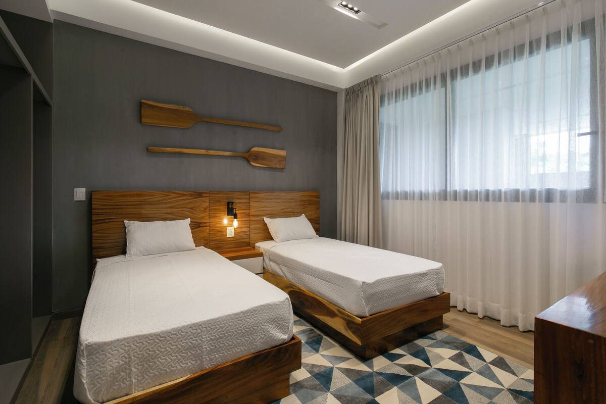 A modern hotel room with two single beds, wooden headboards, a geometric patterned rug, and a large window with sheer curtains.