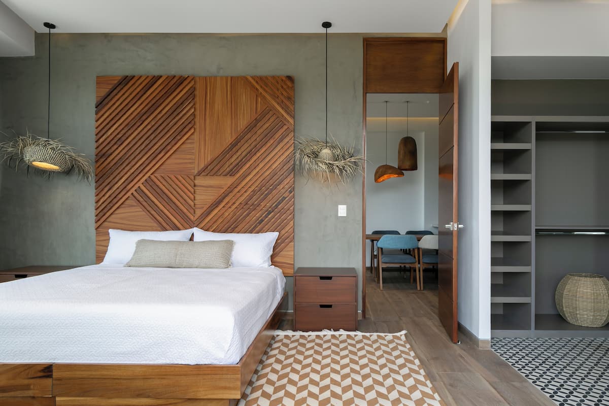 A modern bedroom with a wooden headboard, a bed with white linens, a small wooden nightstand, a woven rattan chair, and a built-in closet with shelves.