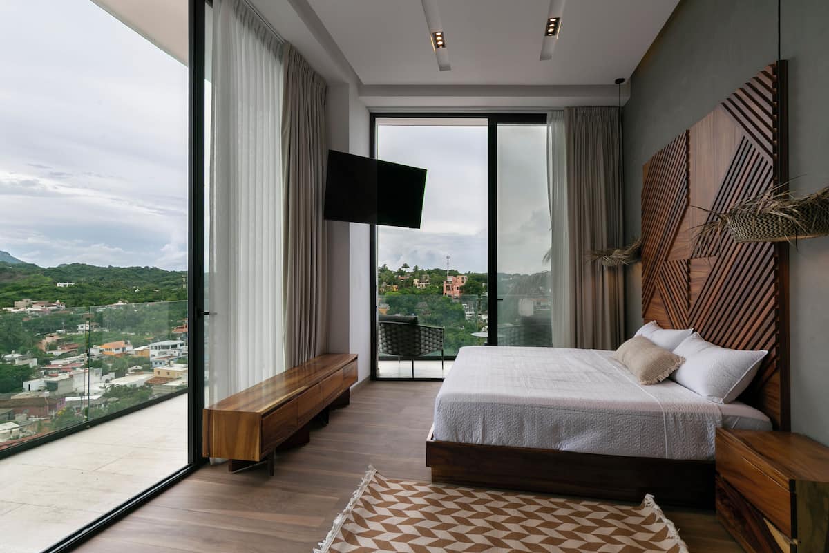 A modern bedroom with a large bed, a bench, and a TV. It has a city view through large windows.