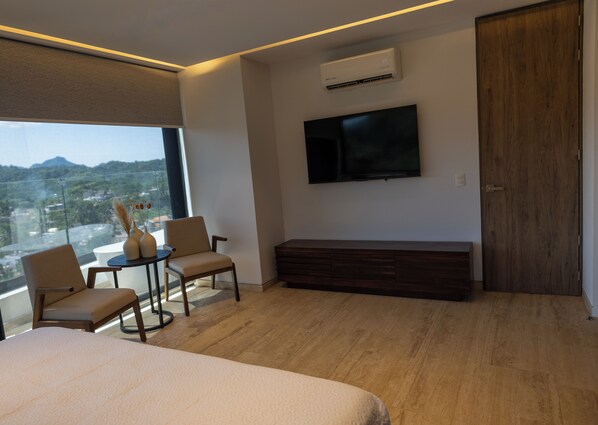 A modern hotel room with a large bed, a seating area with two chairs and a small table, a flat-screen TV mounted on the wall, and a large window offering a city view.