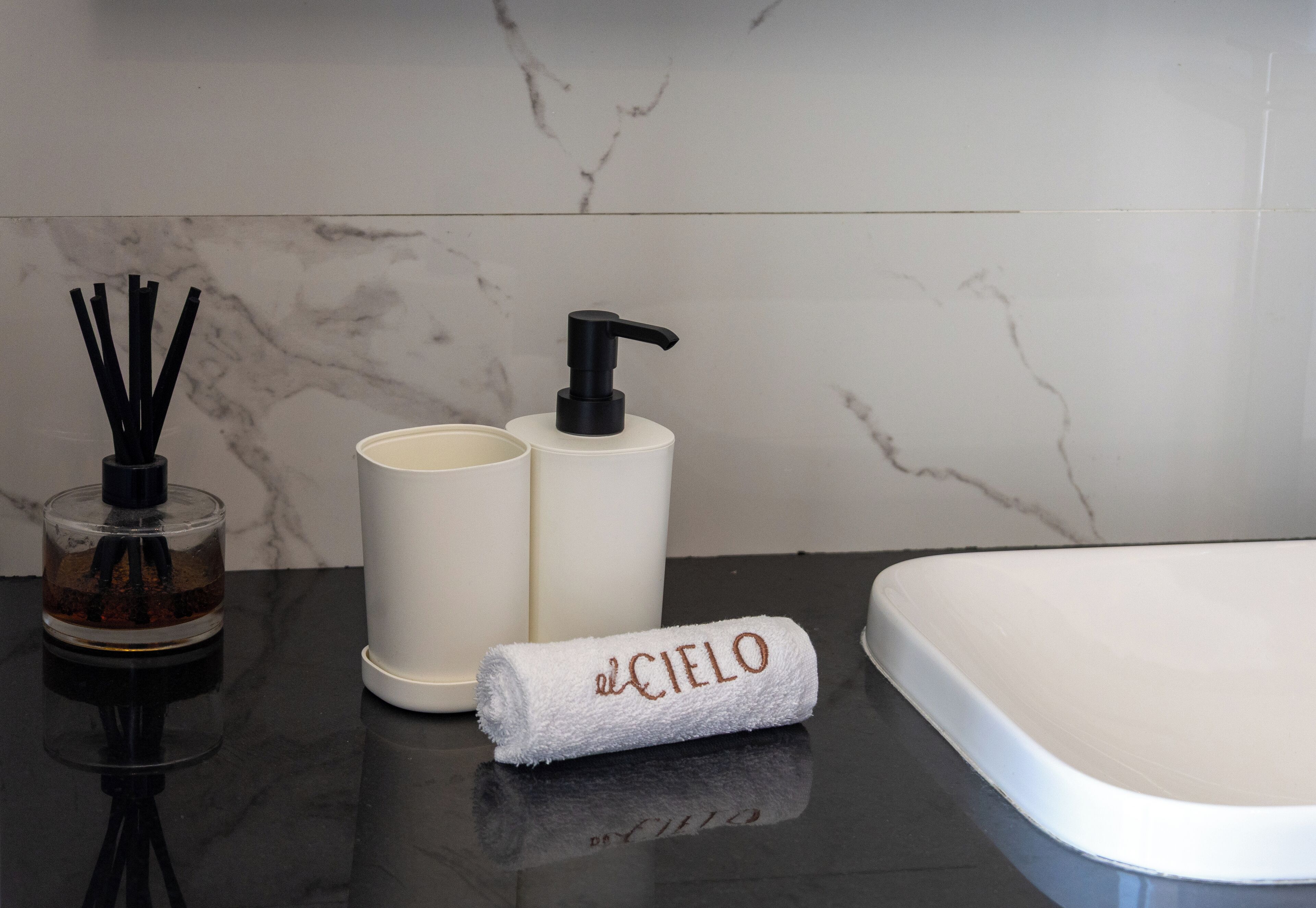A bathroom with a marble wall, a white sink, a black pump bottle, a glass container with black sticks, and a towel with 'el Cielo' embroidered on it.