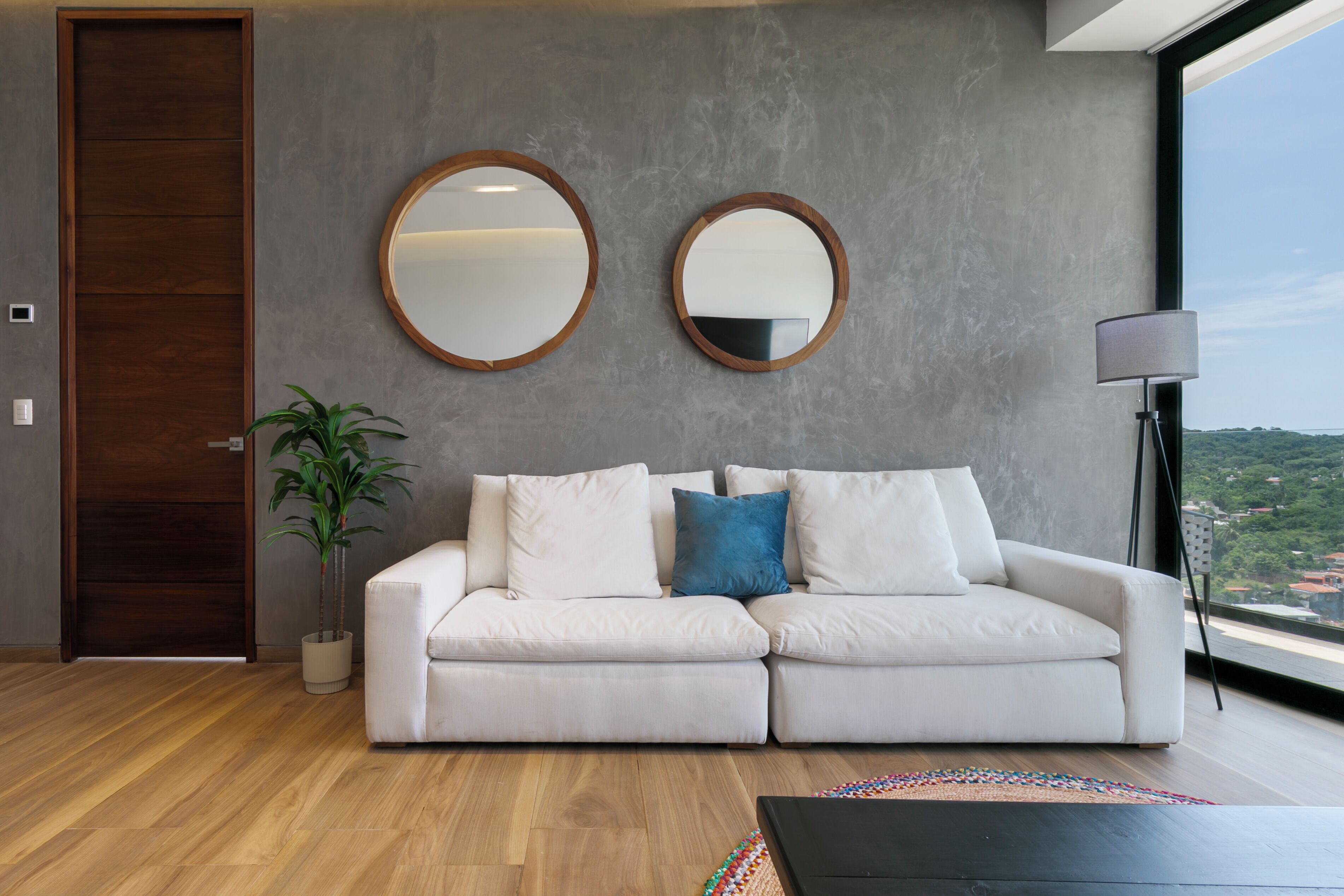 Sala de estar moderna com um sofá branco, dois espelhos redondos, um vaso de planta e vista através de uma janela grande.