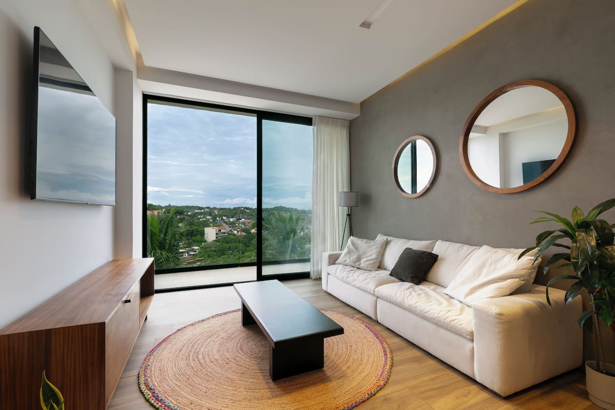 A modern living room with a sofa, a coffee table, a TV, and a large window offering a city view.